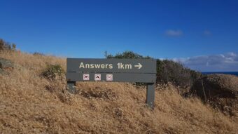 A trail sign reading "Answers 1km" with symbols prohibiting various activities, evoking curiosity about what lies ahead.