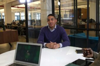 A job candidate in a purple sweater is seated at a table in a modern office, engaged in an interview with a laptop open in front of him