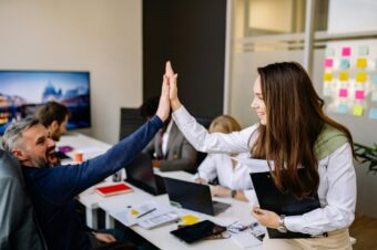 Two colleagues in an office high-five each other, smiling and showing appreciation for their teamwork during a group meeting