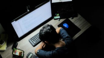 A software developer rests their head on a desk surrounded by multiple screens and devices, illustrating the impact of burnout.