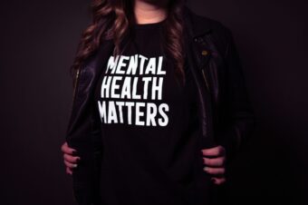 A woman wearing a black t-shirt with the text "Mental Health Matters" stands with her jacket slightly open, showcasing the message. The image is taken in low lighting, emphasizing the text on the shirt. The woman's face is not visible, focusing the attention on the important mental health message.