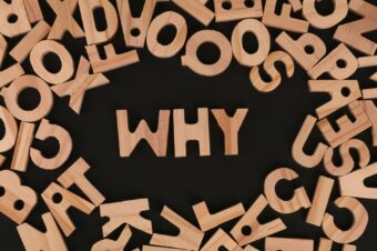 Wooden letters spell out "WHY" amidst scattered letters on a black background, symbolizing the importance of effective questioning.