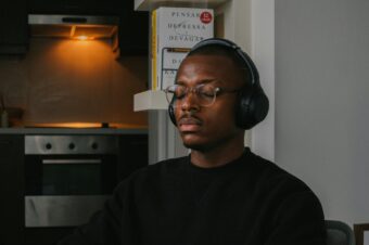 A young man wearing clear glasses and black over-ear headphones is sitting with his eyes closed, appearing to meditate or relax, reflecting a moment of self-awareness. He is in a dimly lit kitchen, with a soft yellow light illuminating the stovetop in the background. A bookshelf with neatly arranged books is next to him, adding to the calm and serene atmosphere.